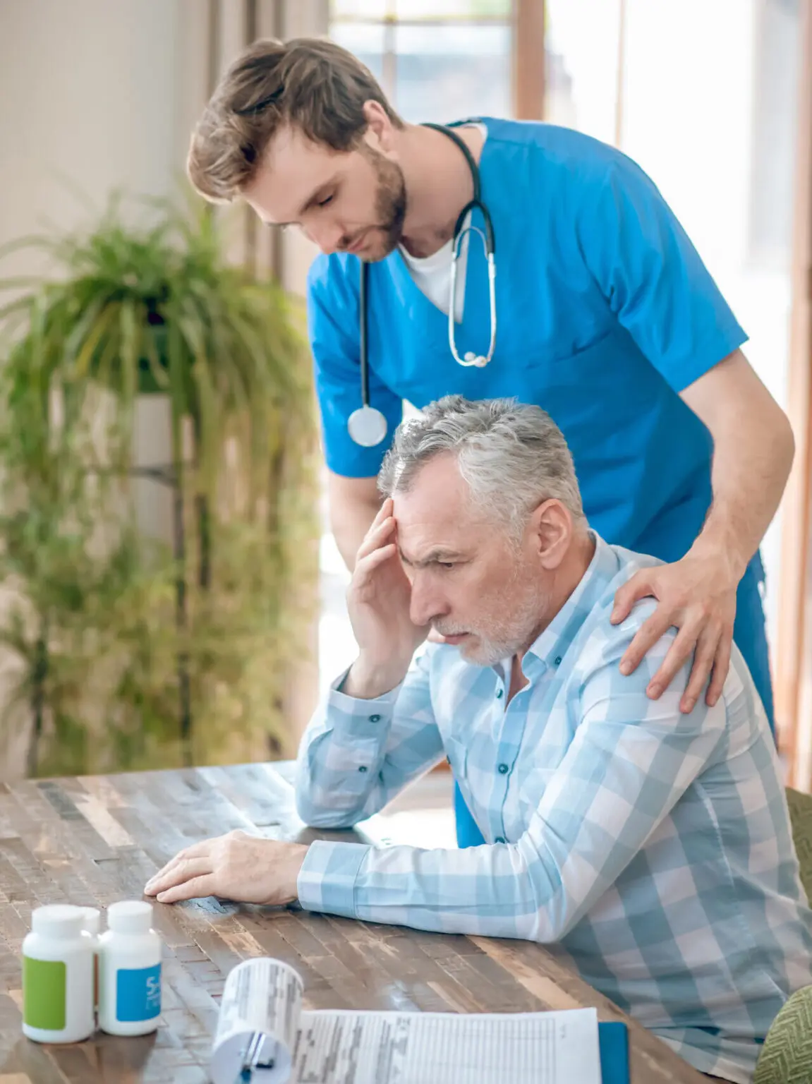 considerate dark haired bearded young caucasian geriatrician with stethoscope leaning sad mature patient edited